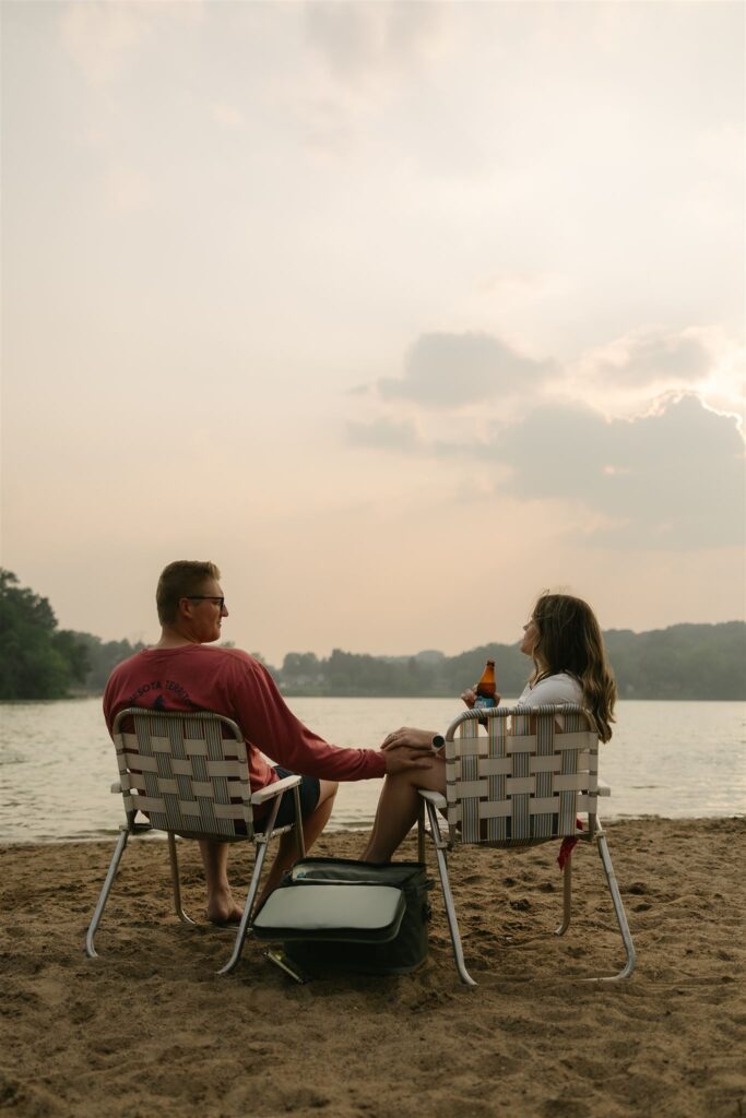 Couple sitting side by side in folding chairs facing the lake, cooler between them.