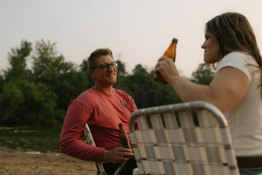 Man smiling and holding a beer while sitting in a beach chair facing woman.