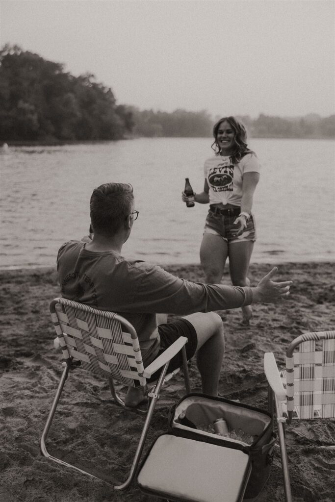 Woman standing on the beach with beer in hand while man sits in chair in foreground.