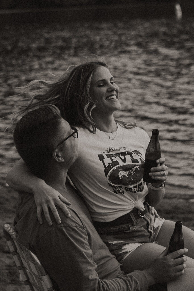 Man sitting on the beach with woman in his lap, both laughing and holding beers for summer engagement photos