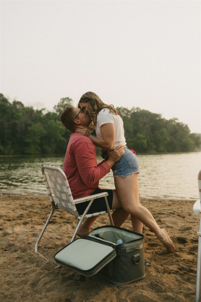 Woman leaning down to kiss man sitting in a folding chair on the beach for summer engagement photos