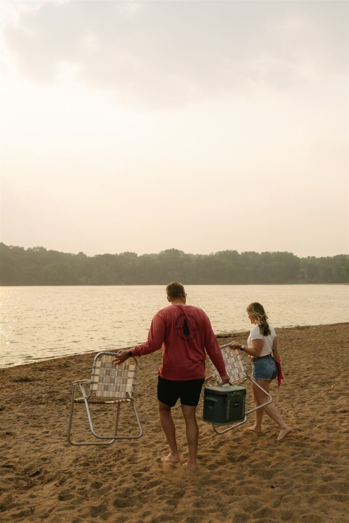 Couple walking across the beach carrying folding chairs and a cooler for summer engagement photos