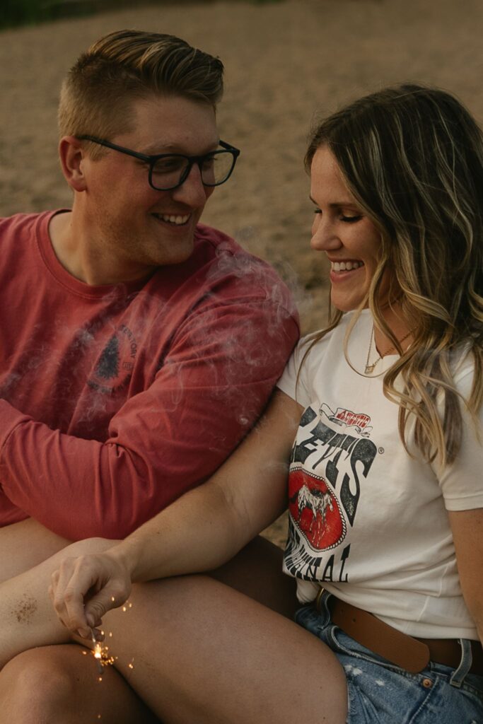 Man holding sparkler while woman smiles beside him for summer engagement photos
