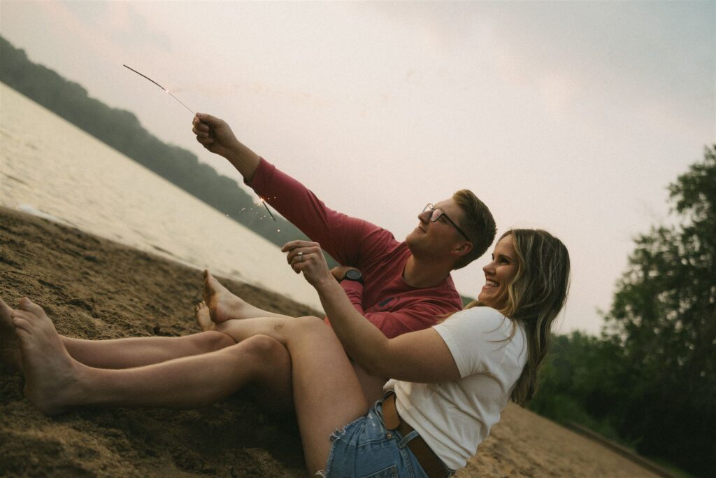 Couple sitting together on the beach, holding sparklers and smiling.