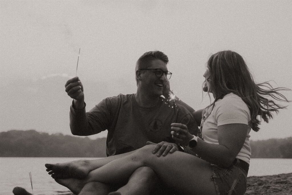 Couple sitting cross-legged together on the sand, each holding a lit sparkler and laughing.