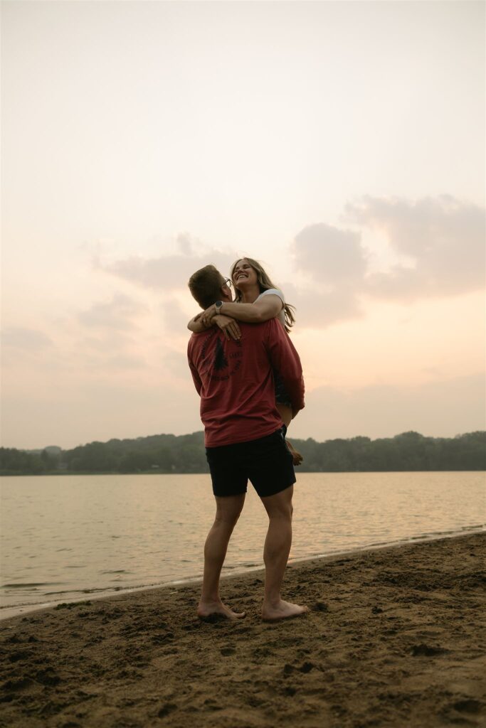 Man lifting woman in his arms on the sandy Minnesota shoreline at sunset.