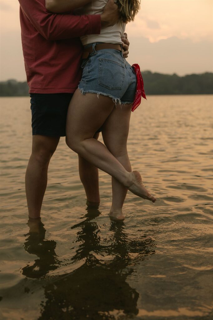 Couple standing ankle-deep in lake water, embracing with her legs wrapped around him.