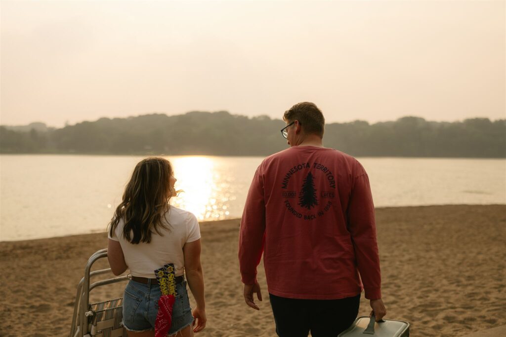 Couple walking on sandy beach toward the water, carrying a cooler and folding chair at sunset during summer engagement photos