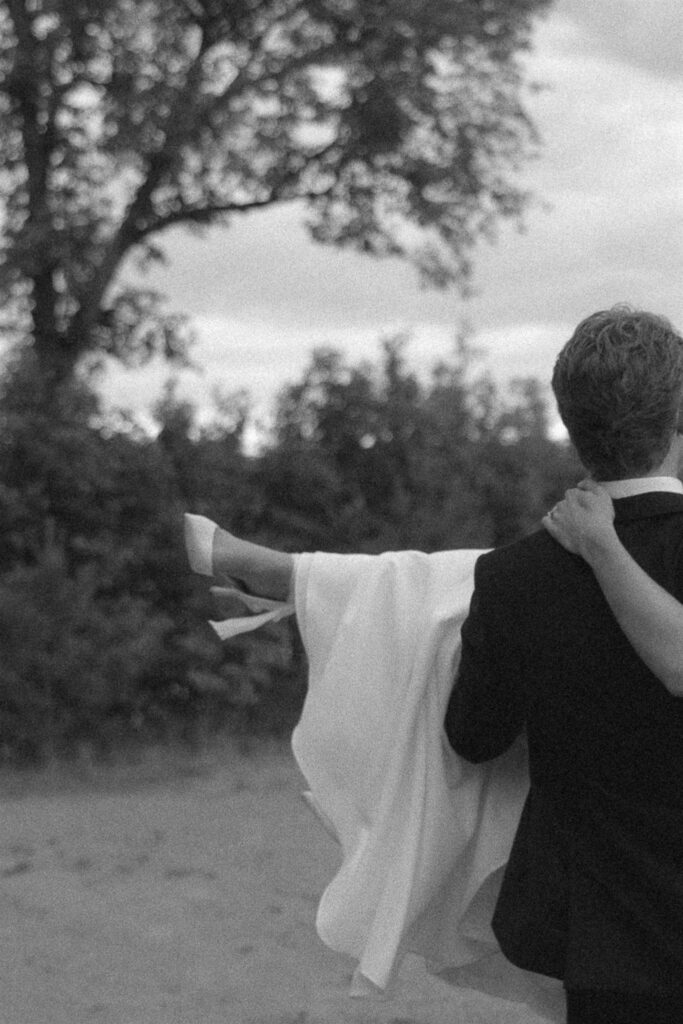 Groom carrying his bride across the grass, her dress and heels lifted, captured in documentary style wedding photos.