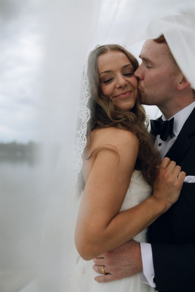 Groom kissing bride’s temple wrapped in her veil, candid lake portrait.