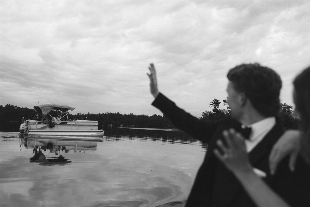 Couple waving to cheering guests from the water, a cinematic lakeside wedding moment.