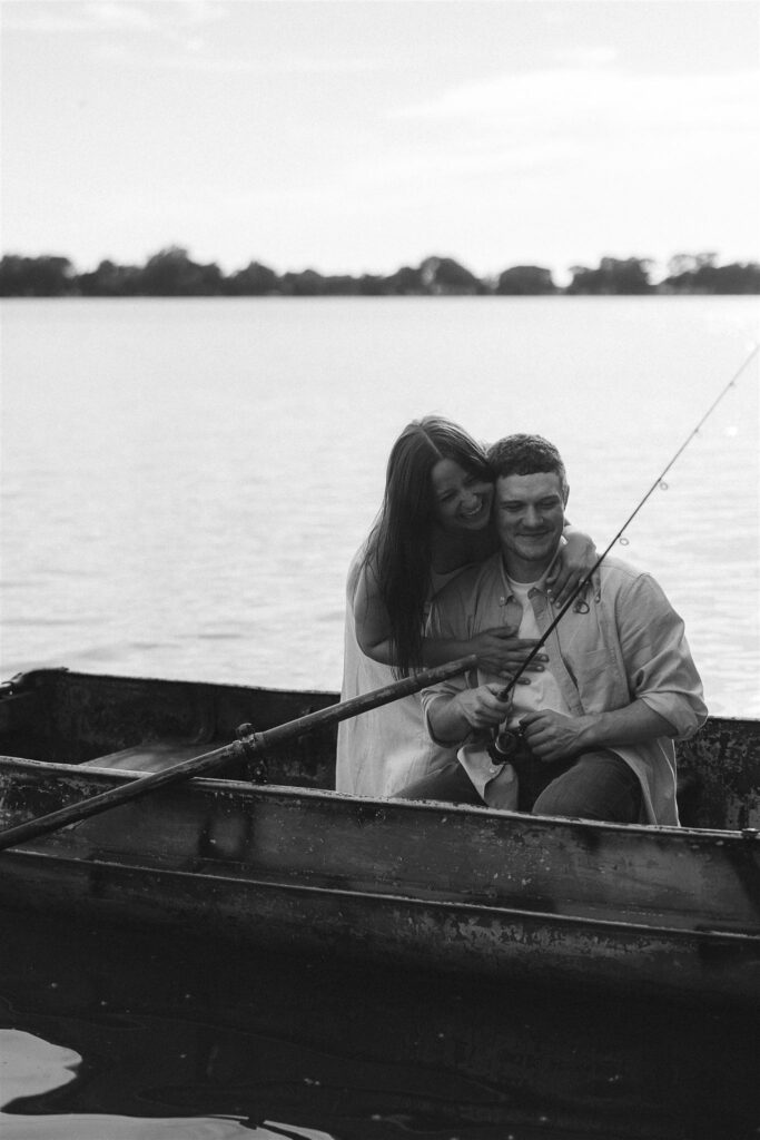 Black and white photo of Sophie and Jake fishing together in the boat, her arms wrapped around him from behind in storytelling engagement photos.