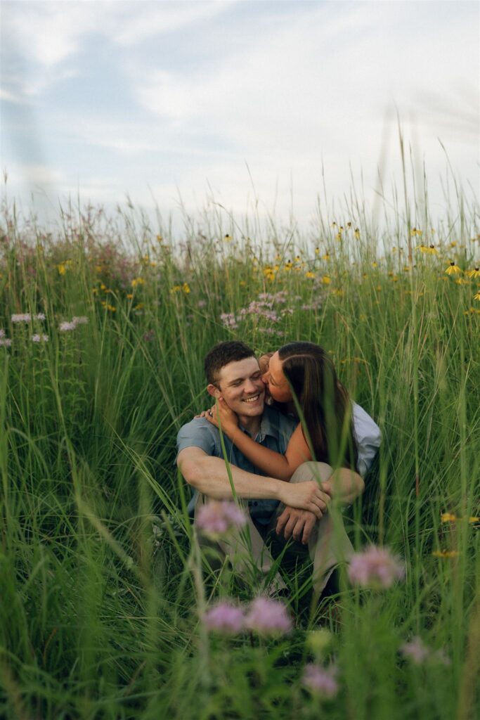 The couple sitting in a wildflower field, Sophie laughing as she kisses Jake’s cheek — pure summer joy.