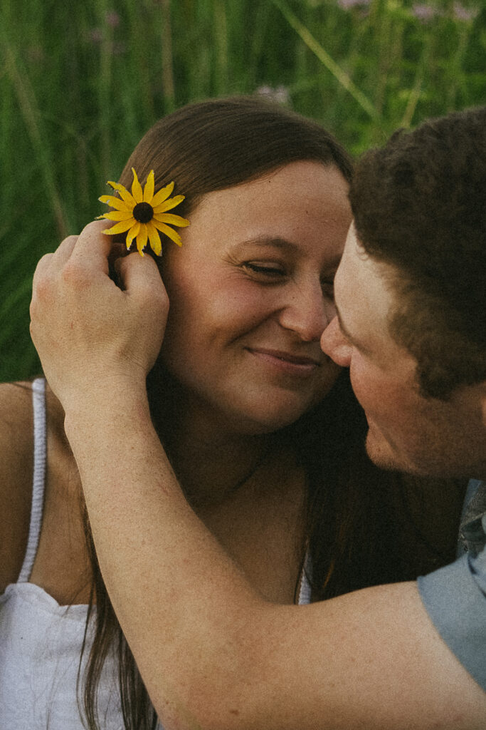 Sophie smiling with a yellow wildflower tucked behind her ear as Jake leans in close, framed by tall grass in storytelling engagement photos.