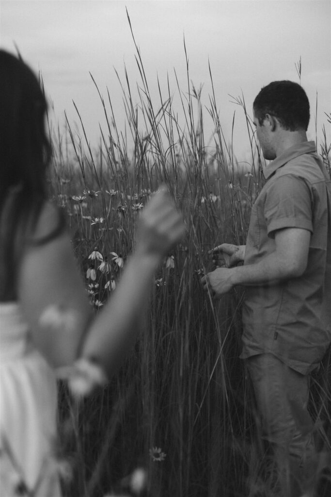 Jake picks wildflowers while Sophie watches from nearby, the two framed in tall grass and fading light, their quiet togetherness captured in a single cinematic photograph.