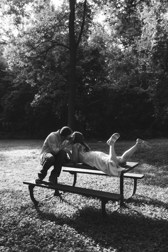 Playful, romantic moment as the couple kisses on top of a picnic table, feet kicked up in the light for their storytelling engagement photos.