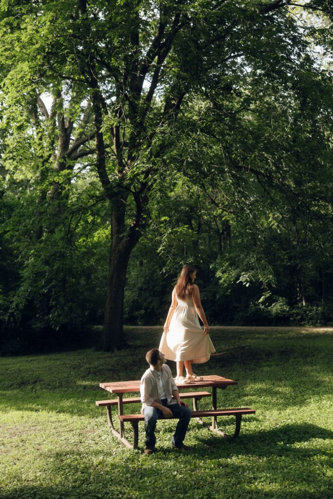 Sophie standing barefoot on a picnic table, dress catching the light, while Jake looks up at her — a cinematic storytelling engagement photos moment.