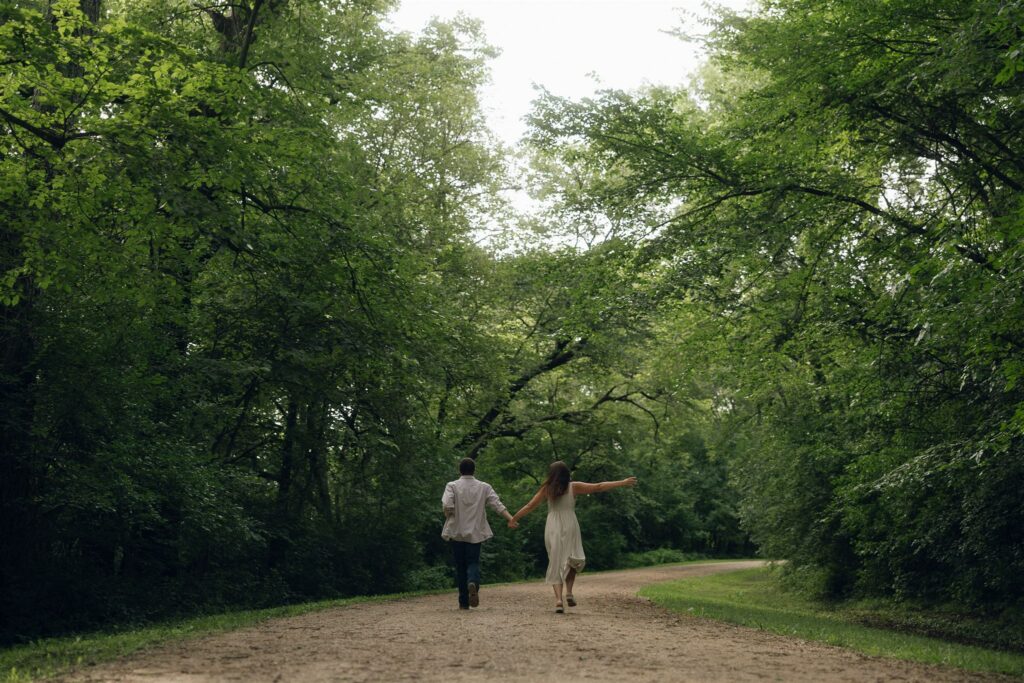 A couple walking hand in hand down a tree-lined dirt road in Minnesota, framed by soft afternoon light and lush summer green in storytelling engagement photos.