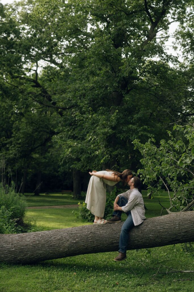 Sophie leaning down to kiss Jake as he sits on a fallen tree, surrounded by greenery and soft summer light.
