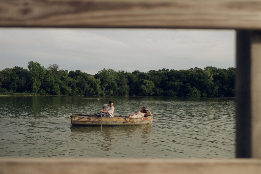 Framed through the dock rails, Sophie and Jake drift across the lake in a weathered rowboat, sunlight soft on the water and trees.