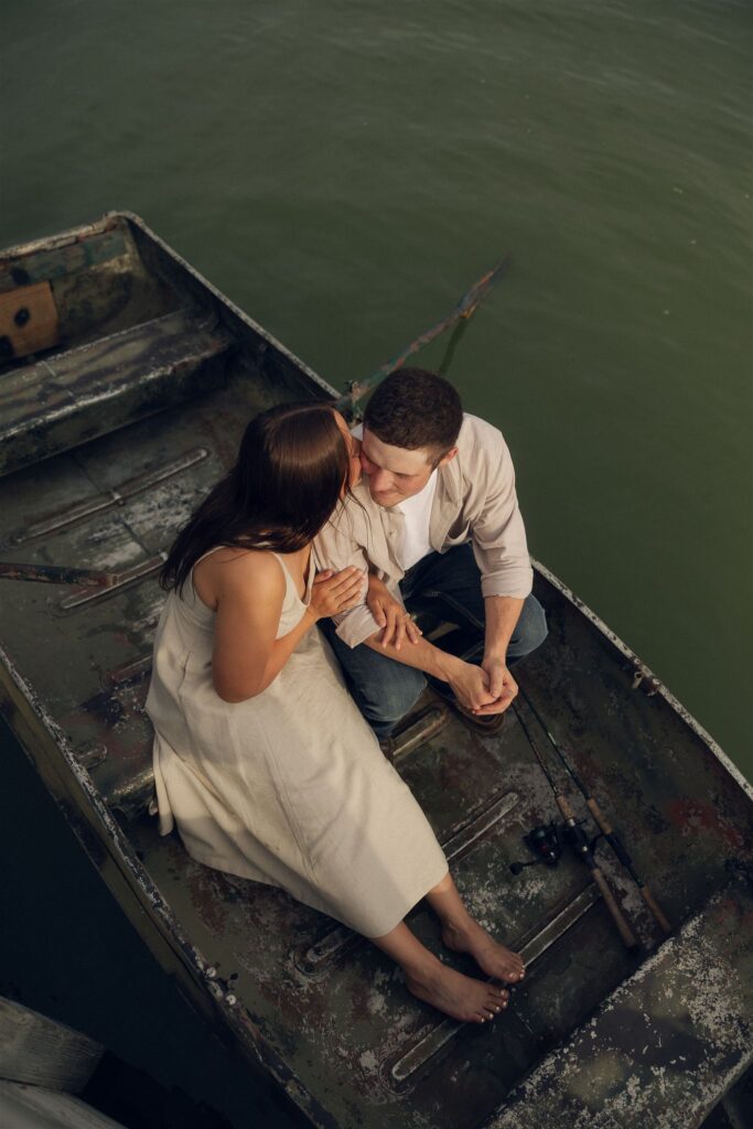 A couple shares a quiet kiss in an old fishing boat drifting across a still Minnesota lake at golden hour, the kind of soft, cinematic moment that defines storytelling engagement photos.