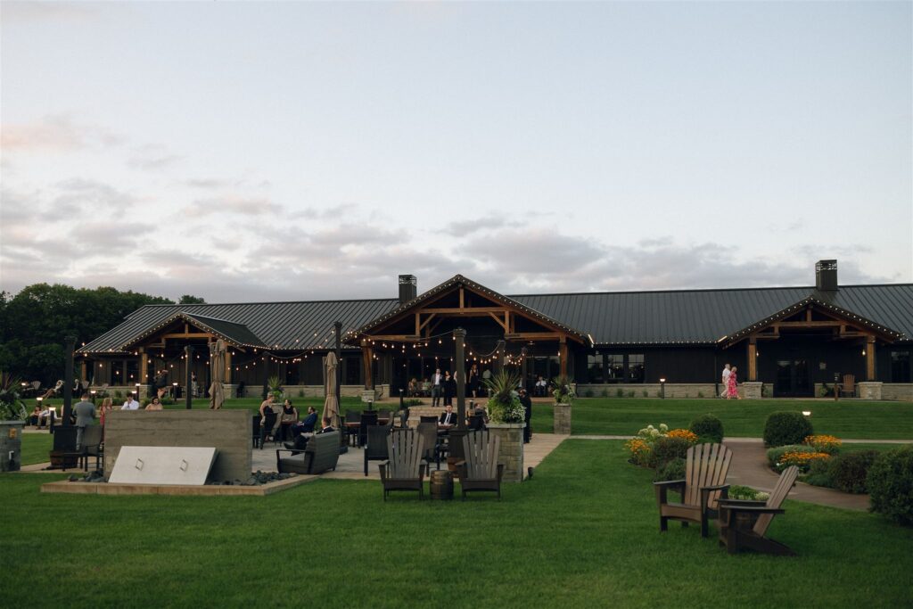 Wide exterior view of the main building at 7 Vines Vineyard and Winery, featuring dark wood architecture, string lights, manicured lawn seating, and guests mingling at sunset at one of the best wedding venues in Minnesota.