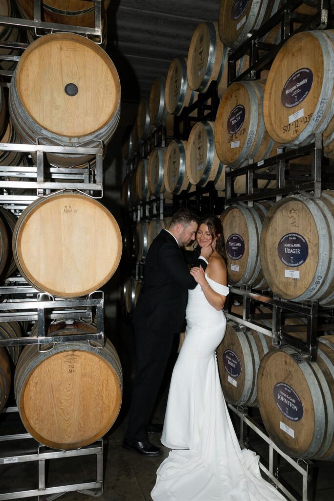 Bride and groom embracing between rows of stacked oak wine barrels inside the barrel room at 7 Vines Vineyard and Winery, with the groom in a black tuxedo leaning in toward the bride in a fitted off-the-shoulder white gown and long train, showcasing one of the most distinctive indoor spaces at a Minnesota wedding venue and highlighting why 7 Vines is considered one of the best wedding venues in Minnesota.