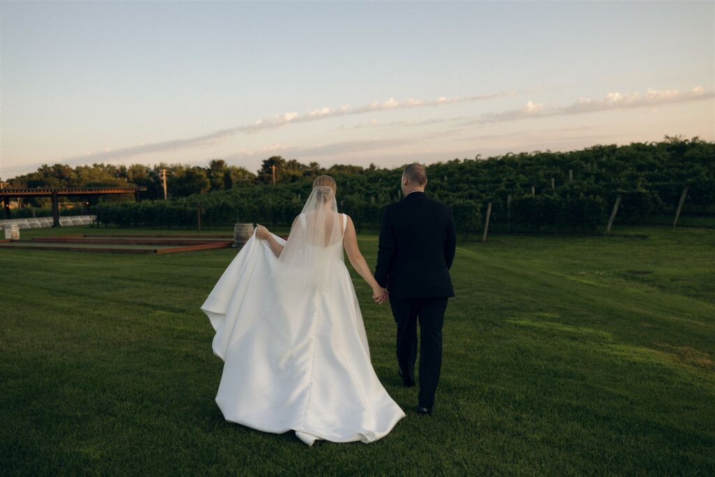 Bride and groom walking away hand in hand across the vineyard lawn at 7 Vines Vineyard and Winery, with grapevines and open sky creating a scenic Minnesota wedding venue backdrop.