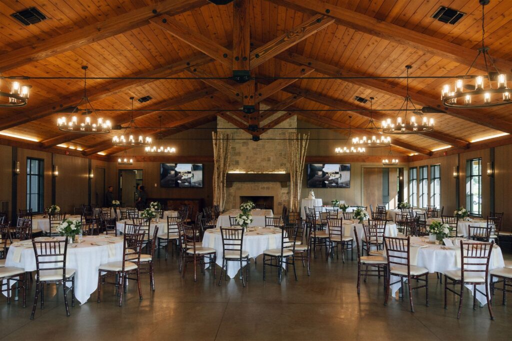 Interior reception space at 7 Vines Vineyard and Winery with exposed wood beams, stone fireplace, round tables, white linens, and chandeliers at one of the best wedding venues in Minnesota.