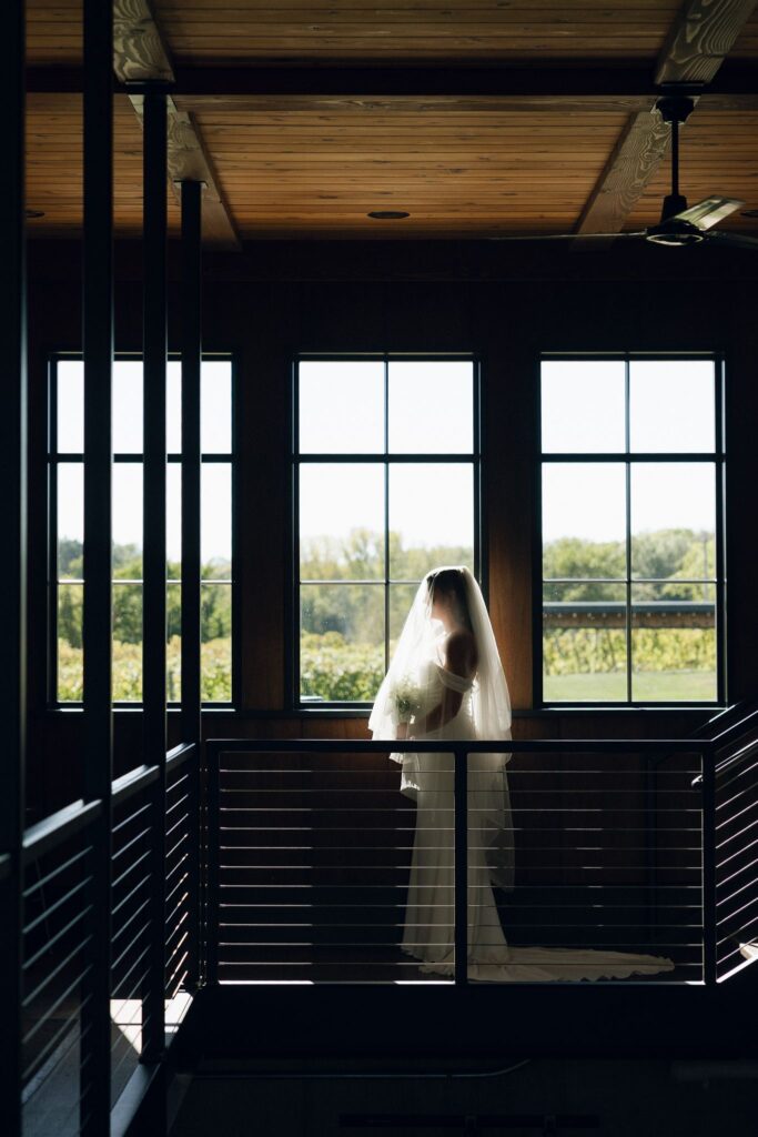Bride standing alone on an indoor balcony at 7 Vines Vineyard and Winery, backlit by large windows overlooking the vineyard at a scenic Minnesota wedding venue.