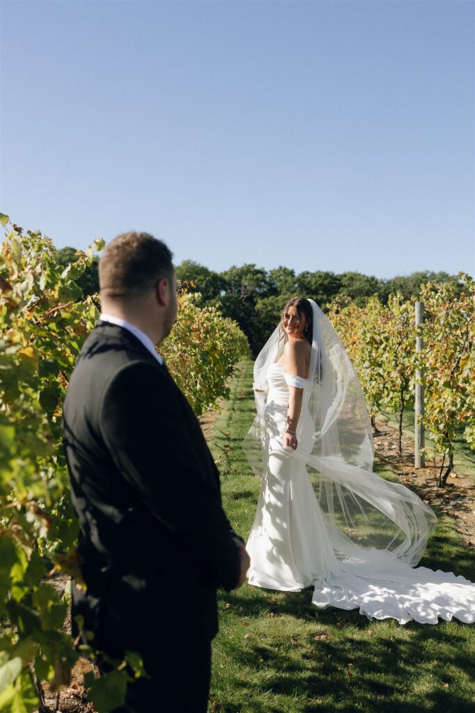 Bride turning back toward groom between vineyard rows at 7 Vines Vineyard and Winery, with veil flowing in the sunlight at one of the best wedding venues in Minnesota.