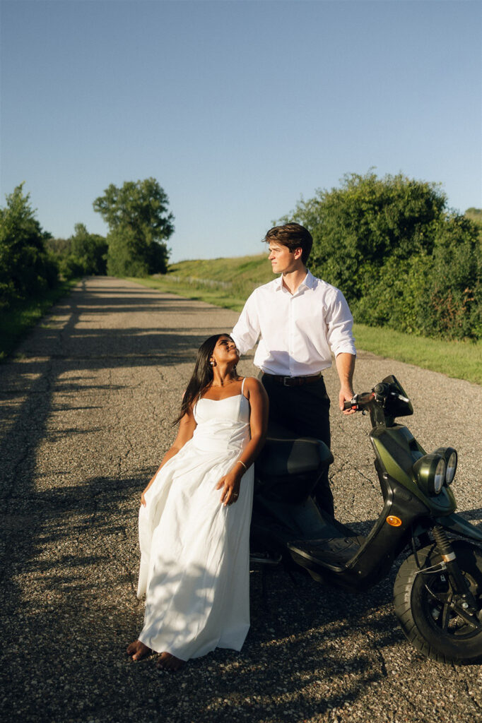 Amira sitting barefoot on the moped while Jake stands beside her on a quiet country road, the kind of classic engagement photos that feel effortless and sun-soaked.