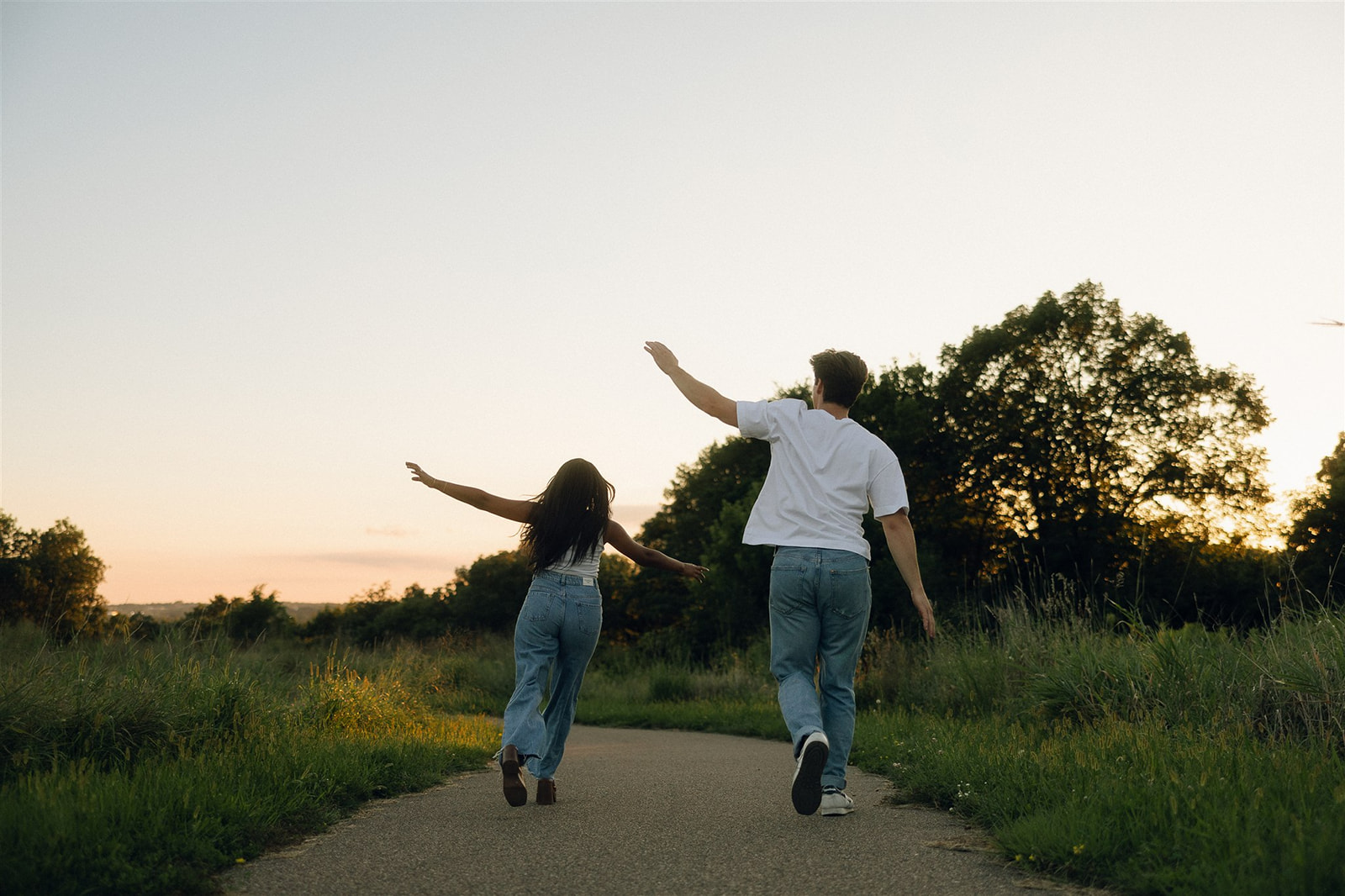 Jake and Amira walking down a paved path through a meadow at sunset, arms stretched wide like kids pretending to be airplanes.