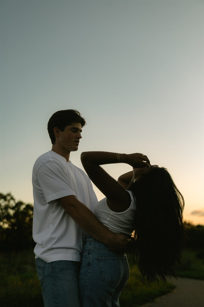 Silhouetted moment of Jake holding Amira at the waist as she tilts her head back laughing, the sky fading into soft dusk behind them.