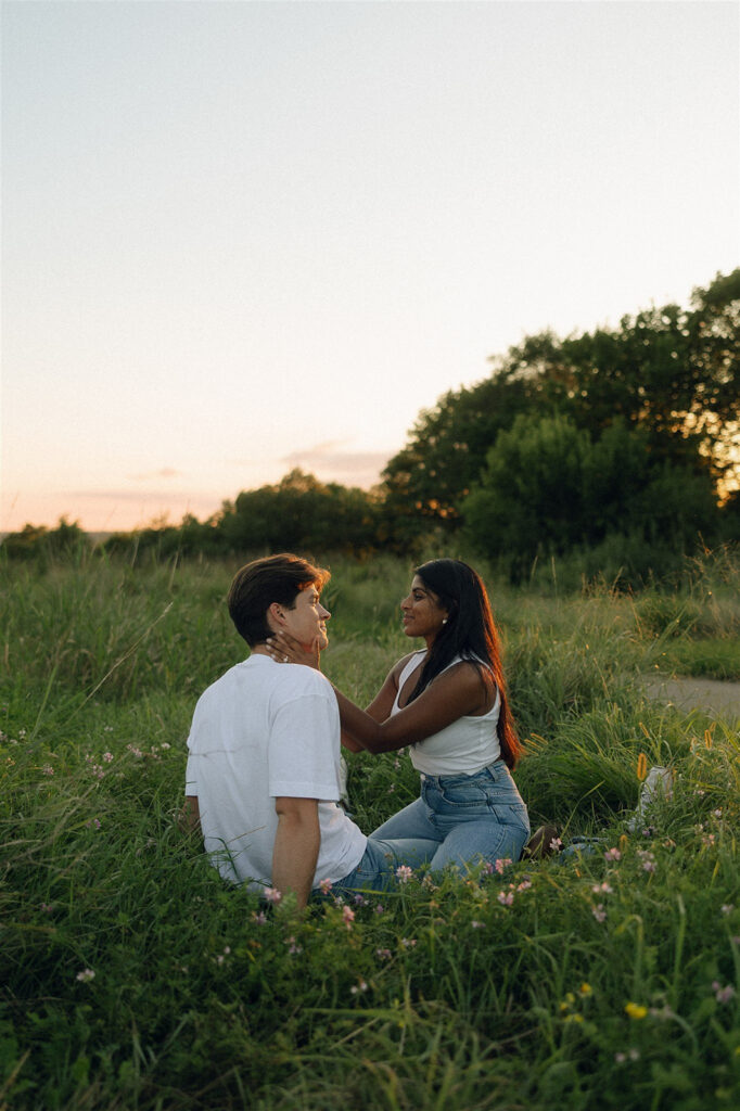 Amira kneeling in the grass in front of Jake, gently holding his face as they talk quietly with the meadow stretching out around them.