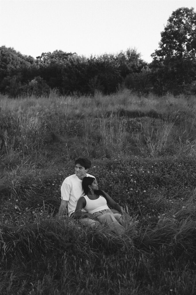 Black and white wide frame of Jake and Amira seated in tall grass, leaning into each other with trees lining the horizon behind them.