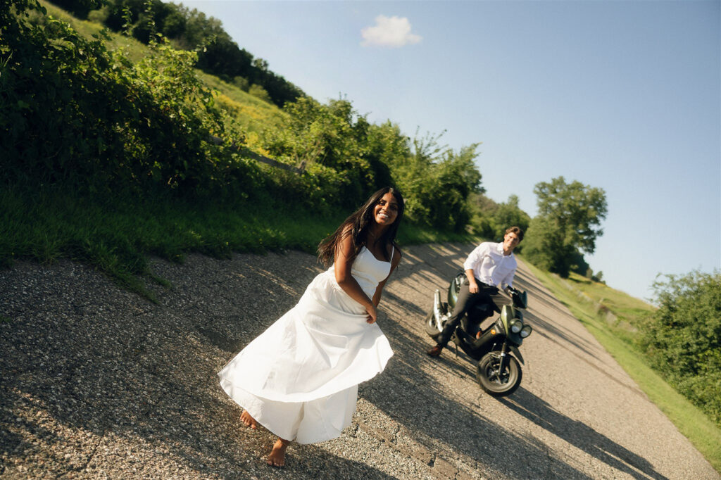 Amira laughing as she moves toward the camera, Jake trailing behind on the moped—an outdoor engagement photo that feels more like a scene than a pose.