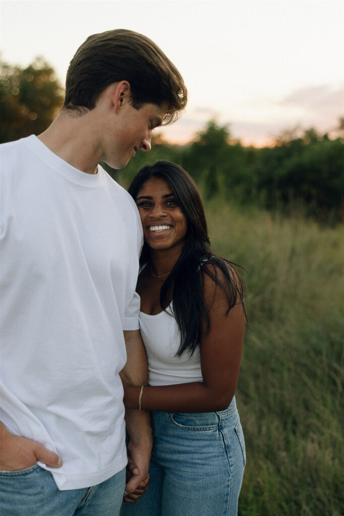 Close-up portrait of the couple standing in a meadow at golden hour, her smiling straight at the camera while he looks down at her with an affectionate grin.