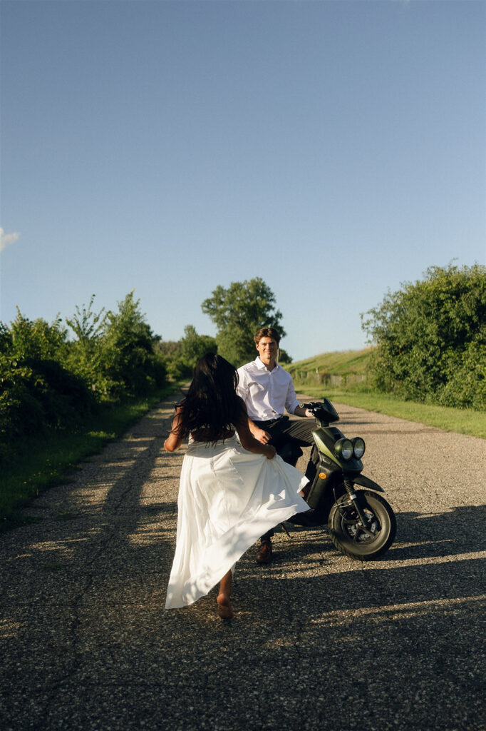 Couple laughing and moving down a rural road with a moped, an effortless take on outdoor engagement photo ideas with modern, cinematic energy.
