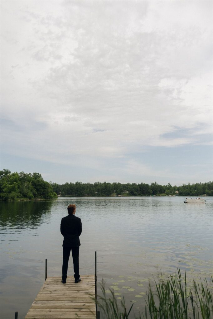 A groom in a black tuxedo stands alone at the end of a narrow wooden dock overlooking a calm Minnesota lake, surrounded by trees and overcast sky at Catalyst by Nature Link, one of the best wedding venues in Minnesota.