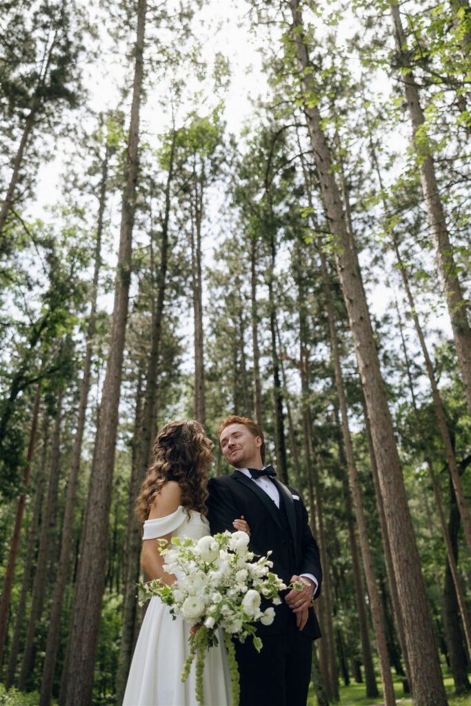 Bride and groom stand together in a tall pine forest at Catalyst by Nature Link, the bride holding a large white floral bouquet while looking up at the groom, showcasing the natural woodland setting of this Minnesota wedding venue.
