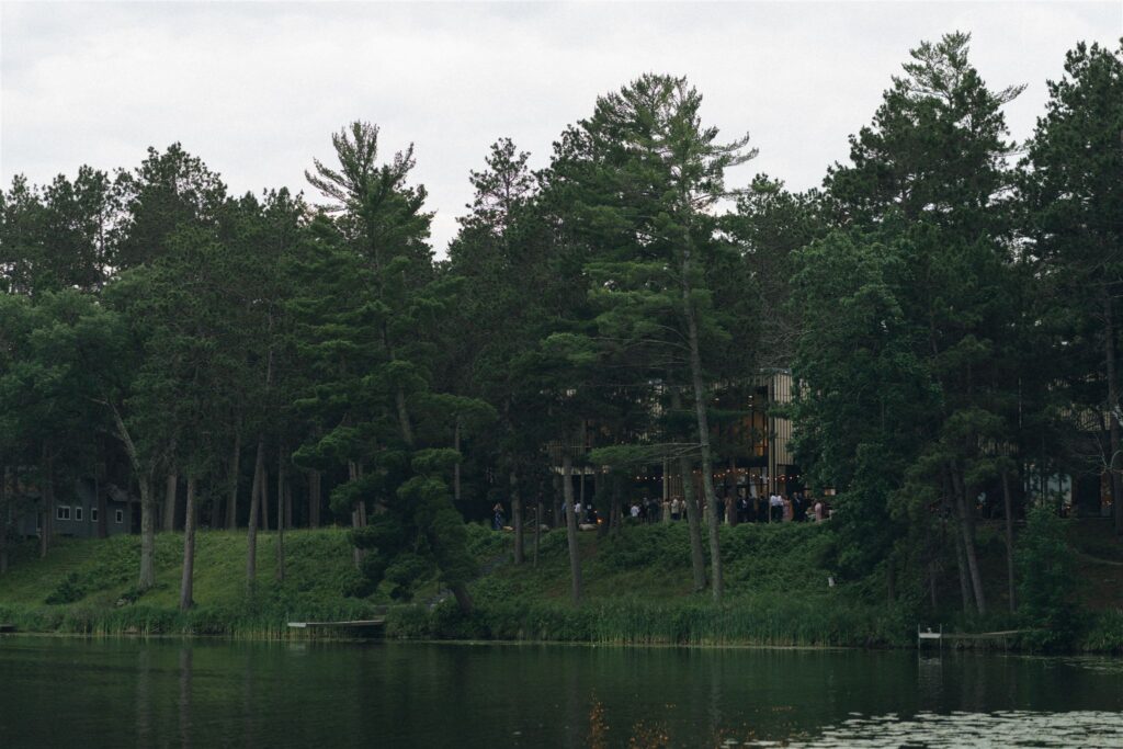 Wide view of Catalyst by Nature Link across the lake, with guests gathered near the modern building nestled among tall pine trees, highlighting the venue’s lakeside forest setting at one of the best wedding venues in Minnesota.