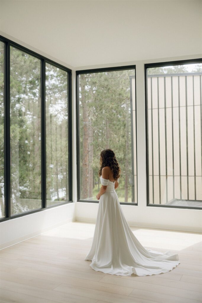 Bride stands alone inside a bright, minimalist room with floor-to-ceiling black-framed windows overlooking pine trees at Catalyst by Nature Link, a modern indoor space within this Minnesota wedding venue.