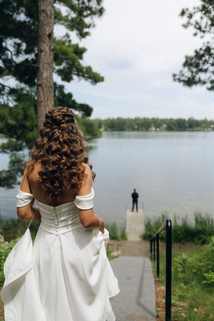 Bride seen from behind walking down a paved path toward a lake, holding her dress while the groom stands waiting at the end of a dock, capturing a quiet first-look moment at Catalyst by Nature Link, one of the best wedding venues in Minnesota.