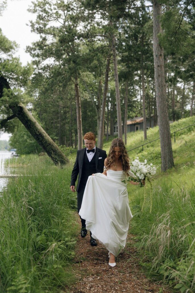 Bride and groom walk together along a narrow forest path at Catalyst by Nature Link, surrounded by tall grass and pine trees, emphasizing the immersive nature setting of this Minnesota wedding venue.