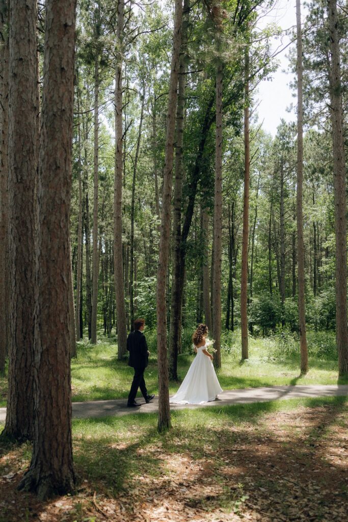 Bride and groom walk side by side on a paved trail through a sunlit pine forest at Catalyst by Nature Link, highlighting the serene woodland paths available at this best wedding venue in Minnesota.