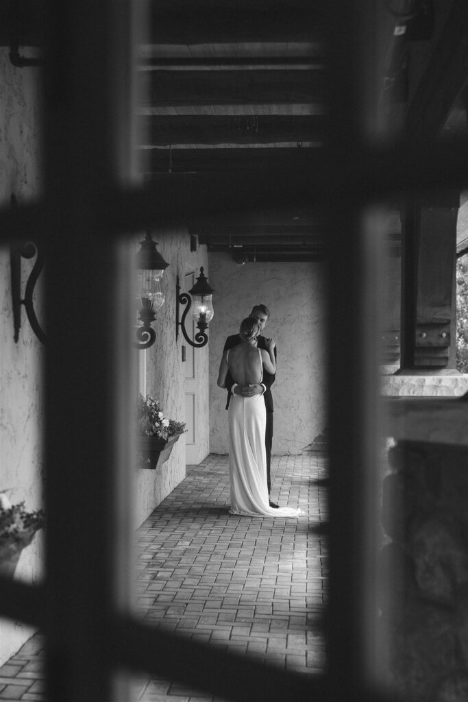 Black-and-white photo of a couple embracing beneath a covered stone walkway at Edward Anne Estate, framed through architectural details.