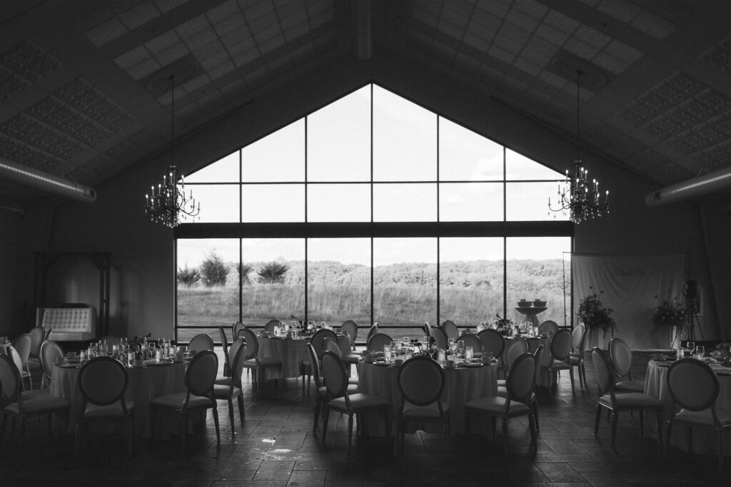 Black-and-white image of the Edward Anne Estate reception space with round tables, chandeliers, and expansive windows overlooking the grounds.