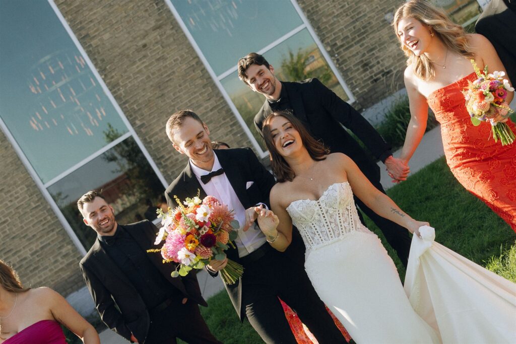 Bride and groom walk hand in hand with their wedding party outside a brick venue, laughing mid-stride as sunlight hits bold dresses and a colorful wedding bouquet.