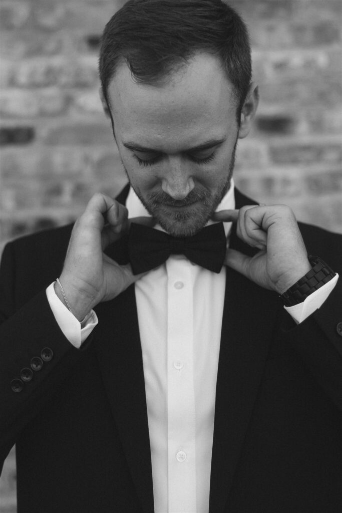 Black-and-white portrait of a groom adjusting his bow tie, head tilted down in a quiet getting-ready moment against a brick wall.